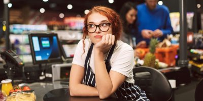 Žena s brýlemi sedí znuděně v pokladně obchodu