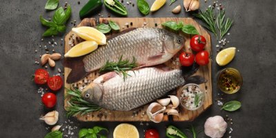 2 kapři na dřevěném prkénku, kolem česnek, rajčata, zelenina