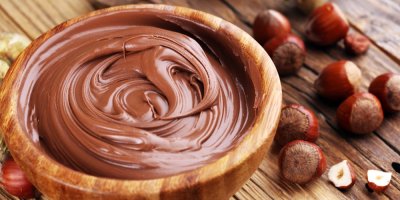 Hnědá pomazánka v dřevěné misce na dřevěném stole, na němž jsou také lískové oříšky