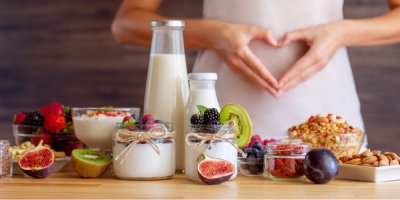 Zdravé potraviny v popředí, v pozadí žena dělá rukama srdce na břichu