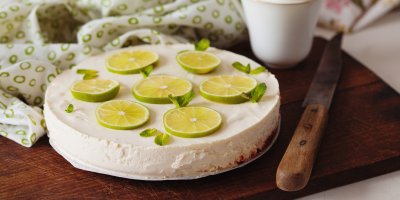 Limetkový cheesecake v celku, jsou na něm kolečka limetek a vedle nůž, hrnek a utěrka