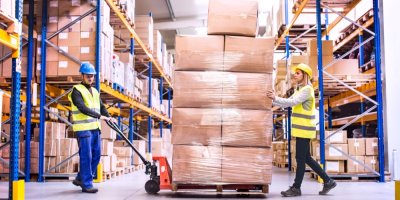 Muž a žena pracující ve skladu přepravují na vozíku náklad krabic obalených plastovou fólií.