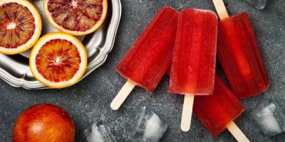 Několik červených nanuků z Aperolu vyskládaných na stole vedle kovového tácku s plátky pomerančů