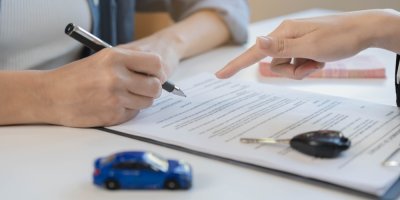 Žena podepisuje dokument, ruka úřednice ukazuje na místo podpisu, na stole jsou klíče od auta hračka auta