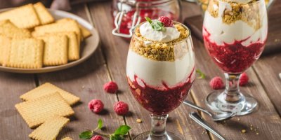 2 skleničky s nepečeným malinovým cheesecakem. Kolem skleniček maliny a talíř s máslovými sušenkami