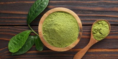 Dřevěná miska plná matcha prášku, vedle ní je dřevěná lžička s matchou a zelené listy.