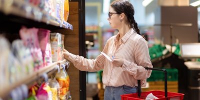 Žena v košili s nákupním košíkem stojí u regálu a vybírá si výrobek
