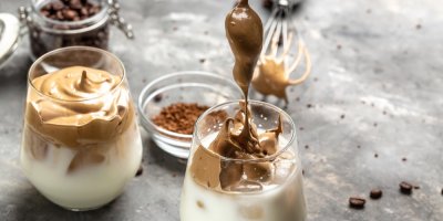 Dvě sklenky s mlékem, ledem a našlehanou kávovou pěnou, okolo jsou rozsypaná kávová zrna