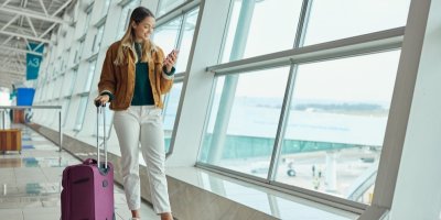 Žena stojí s kufrem na letišti a dívá se na mobil