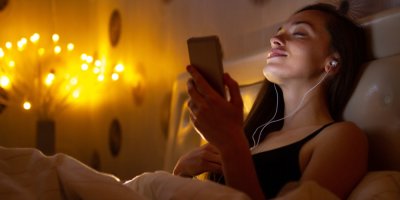 Žena sedí v posteli a má v uších sluchátka, v ruce drží telefon