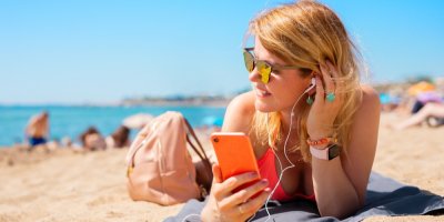 Mladá žena leží na pláží a poslouchá hudbu ze sluchátek, připojených do telefonu, který drží v ruce