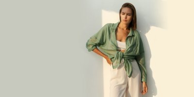 Žena se opírá o stěnu, na sobě má bílé plátěné kalhoty a zelenou volně uvázanou lněnou košili.