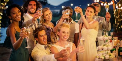 Svatebčané a novomanželé si připíjejí šampaňským, jedna z hostek má bílé šaty