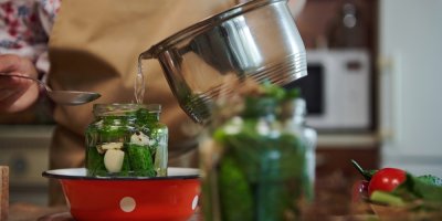 Žena v zástěře lije lák do připravené sklenice s okurkami, kořením a zeleninou.
