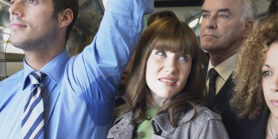 Žena stojí uprostřed davu v hromadné dopravě, vedle ní stojí muž se zpoceným podpažím, který se rukou drží madla
