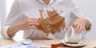 Žena se drží za košili s kávovou skvrnou, na stole rozlitý hrnek kávy