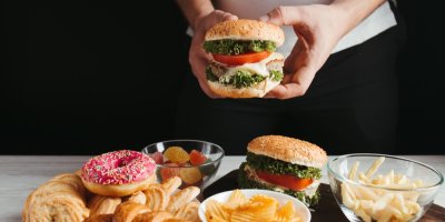 Ruce drží hamburger, na stole je sladké a slané pečivo, bonbóny, chipsy