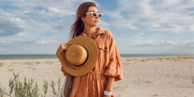 Mladá žena ve volných lněných šatech, slunečních brýlích a s kloboukem v ruce