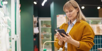 Žena s nákupním košíkem v rukou si v obchodě prohlíží něco v telefonu.