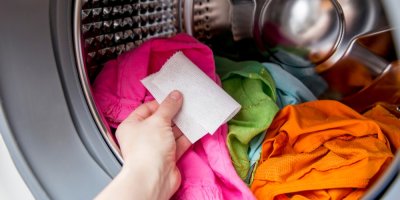 Ruka vkládá do pračky plné barevného prádla ubrousek pohlcující obarvení.