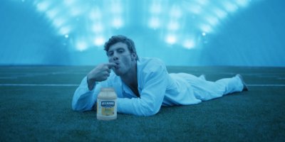 Muž leží břichem na trávě, olizuje si ukazováček, před ním je sklenice majonézy Hellmann's