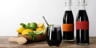 Colový nápoj ve skleničce s brčkem, v pozadí dvě plné lahve a mísa s citrusy