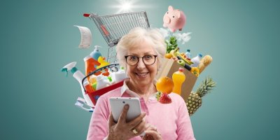 Seniorka drží v ruce telefon, v pozadí nákupní vozík a zboží