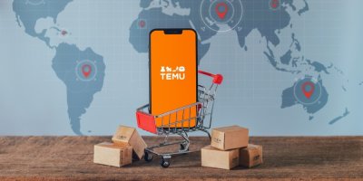 Telefon s logem Temu v miniaturním nákupním vozíku, v pozadí mapa světa