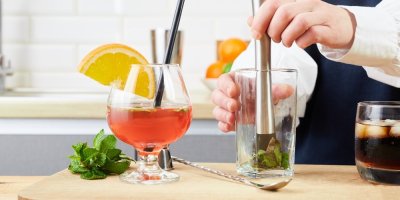 Žena drtí bylinky ve sklenici, vedle jsou sklenice s míchanými nápoji