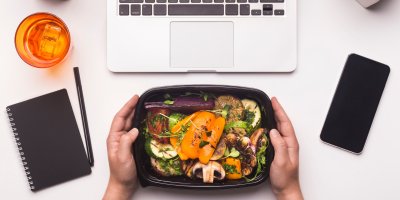 Ruce držící krabičku s jídlem na pracovním stole s notebookem, sešitem, skleničkou a telefonem 