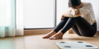 Žena sedí smutně v pozadí, v popředí je váha