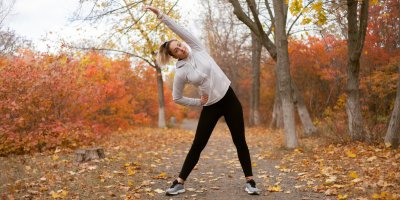 Žena v legínách a mikině stojí na cestě plné opadaných listů a protahuje se