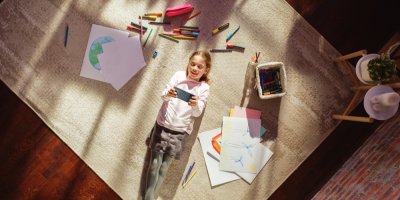 Dívka leží na koberci a drží telefon, kolem jsou pastelky a výkresy