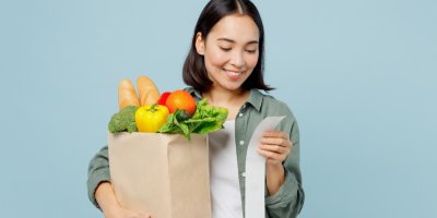 Mladá žena drží v ruce tašku s nákupem a ve druhé ruce účtenku, usmívá se 