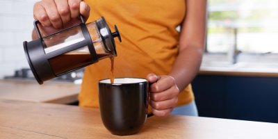 Žena nalévá z french pressu kávu do hnědého hrnku.