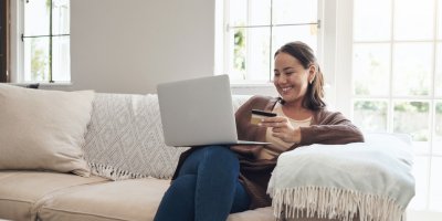 Žena sedí na gauči, na klíně má notebook, v ruce platební kartu a usmívá se