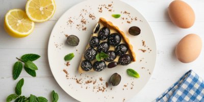 Koláč posypaný černými hrozny na bílém talíři, kolem citrony a vejce