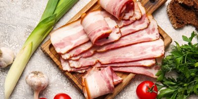 Plátky slaniny na dřevěném prkénku, kolem zelenina