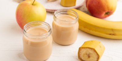 Dvě dětské přesnídávky ve skleničce, rozkrojený banán a jablka