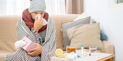 Nemocný člověk zabalený v dece smrká a na stole má léky s čajem