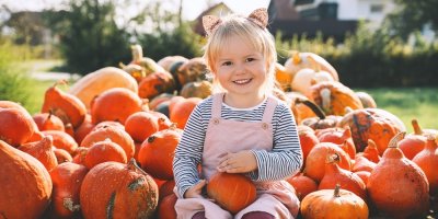 Malá holčička sedí na korbě plné dýní a jednu drží v ruce