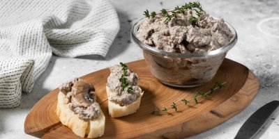 Houbová paštika ve skleněné misce a namazaná na bagetku, na dřevěném prkénku