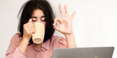 Rozespalá žena sedící za notebookem pije kávu a gestem ruky ukazuje, že je vše v pořádku