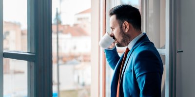 Muž v obleku se dívá z okna a pije nápoj z bílého hrnku