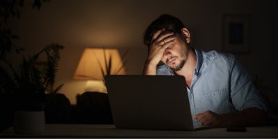 Muž sedí v tmavé místnosti u laptopu s hlavou v dlaních.