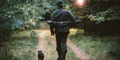 Myslivec má na zádech loveckou pušku a se svým psem jde po lesní cestě