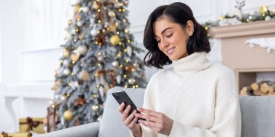 Žena sedící u vánočního stromečku si s úsměvem prohlíží chytrý telefon.