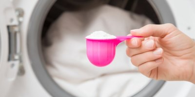 Ženská ruka drží odměrku plnou pracího prášku, v pozadí je plná pračka