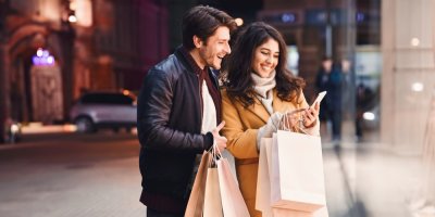 Muž a žena se s nákupními taškami v rukou s úsměvem dívají do chytrého telefonu.