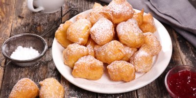 Kousky smaženého těsta na talíři, posypané moučkovým cukrem, vedle mistička s džemem